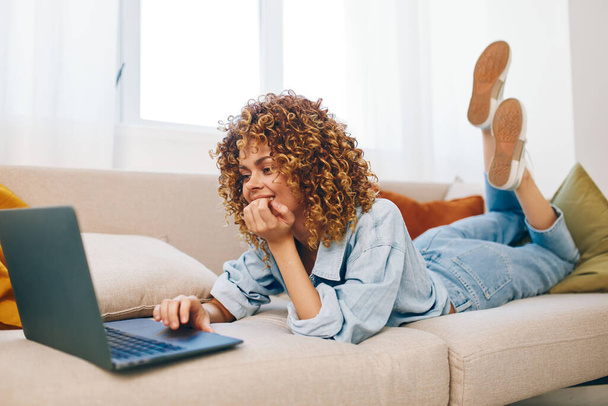 Усміхнена жінка працює на ноутбуці в затишному домашньому офісі, оточеному сучасними технологіями та відпочинку на дивані - Фото, зображення