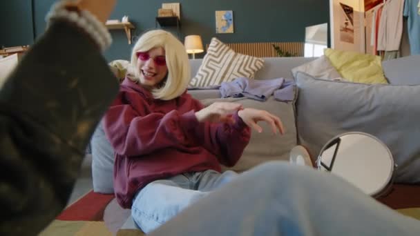 Schnappschuss eines kaukasischen Teenagers mit Zerebralparese mit blonder Perücke und rosafarbener Y2k-Brille, der im modernen Wohnzimmer auf dem Boden sitzt, tanzt und mit einem Freund plaudert und drinnen chillt - Filmmaterial, Video