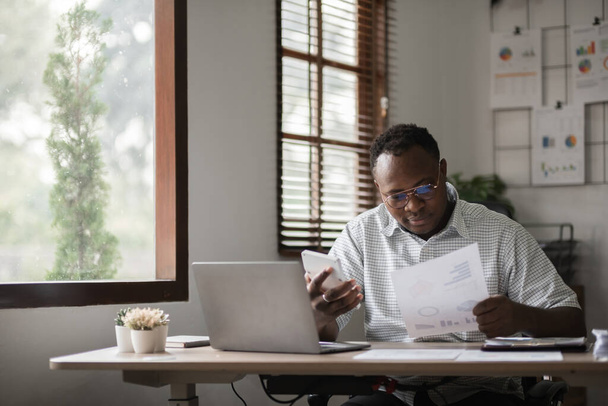 Afrikkalainen amerikkalainen liikemies työskentelee paperitöiden parissa, etsii tietoja kannettavalla tietokoneella kotitoimistossa. - Valokuva, kuva