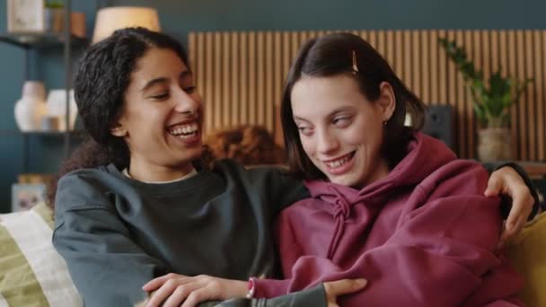 Střední pomalý portrét dvou různorodých veselých dospívajících dívek objímajících a smějících se na kameru sedících na gauči v jasném moderním obývacím pokoji - Záběry, video