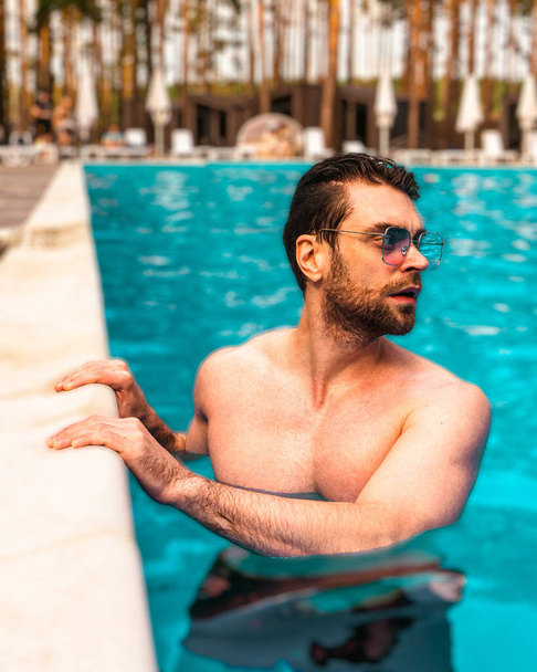 Tipo barbudo en gafas de sol mirando hacia otro lado mientras descansa en la piscina al aire libre. Verano, vacaciones, concepto de estilo de vida - Foto, Imagen