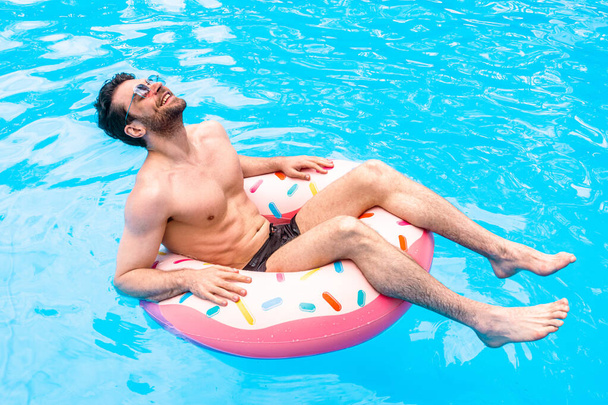 Glücklicher junger Mann schließt die Augen, während er sich im Swimmingpool auf dem aufblasbaren Ring ausruht. Sommerzeit, Urlaub, Lifestylekonzept - Foto, Bild