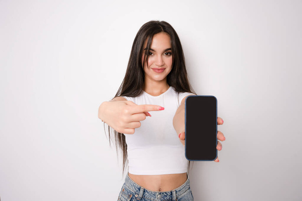Lächelnde junge Frau zeigt leeren Smartphone-Bildschirm und zeigt vereinzelt auf weißen Hintergrund. - Foto, Bild