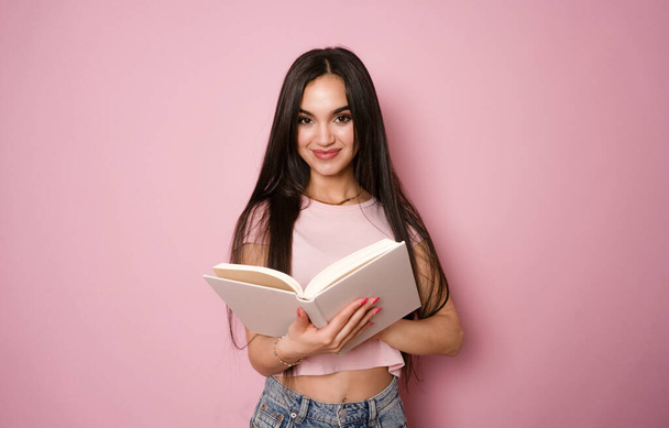 Молода жінка посміхається, тримає відкриту книгу, ізольовано на фоні рожевого кольору - Фото, зображення