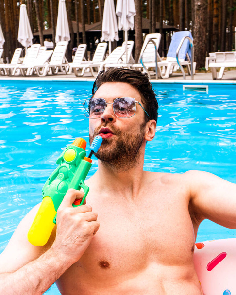 Atlético joven sosteniendo pistola de agua de juguete mientras disfruta de su fin de semana en la piscina al aire libre. Verano, vacaciones, concepto de estilo de vida - Foto, imagen