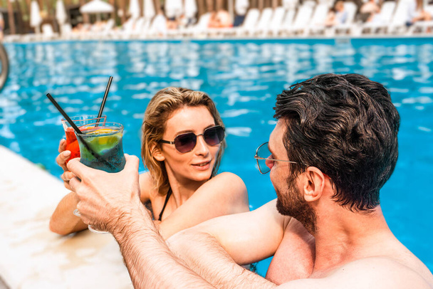 Glimlachende blonde jongedame en haar metgezel klinkende glazen met cocktails in een zwembad van het vakantieoord. Vrije tijd en zomervakantie concept - Foto, afbeelding
