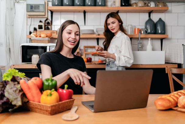 Twee jonge vrouw leren en kijken naar voedsel recept tutorial op laptop samen tijdens het koken van gezonde maaltijd met genieten in de moderne keuken thuis. - Foto, afbeelding