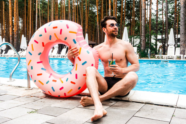 Улыбающийся парень в солнечных очках наслаждается коктейлем, держа надувное кольцо возле бассейна. Лето, праздники, образ жизни - Фото, изображение