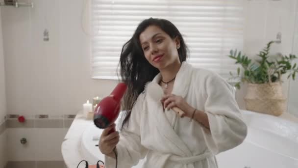 Средний снимок молодой белой девушки, которая делает прическу с феном и расческой в ванной комнате - Кадры, видео