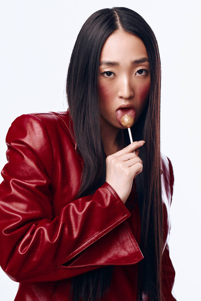 Elegante mujer asiática en chaqueta de cuero rojo Holding Lollipop en la boca al aire libre en el entorno urbano - Foto, imagen