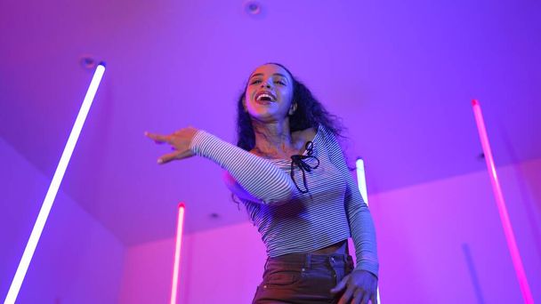 Tieffliegerkamera einer hispanischen Tänzerin, die in die Kamera blickt und zur Popmusik der Stadt tanzt. Professionelle attraktive Hipster bewegen sich zu Hip-Hop-Songs mit lila LED- oder Neonlicht. Stilvolles Tuch. Regalisation. - Foto, Bild