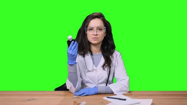 医薬品のボトルを示す保護手袋で制服を着た女性医師は,緑色の隔離された背景に机に座っています. 医療・医療コンセプト - 写真・画像