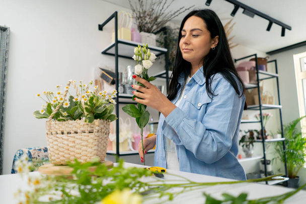 in een bloemenwinkel een meisje bij een witte tafel verzamelt een bloemstuk een boeket in een mand - Foto, afbeelding