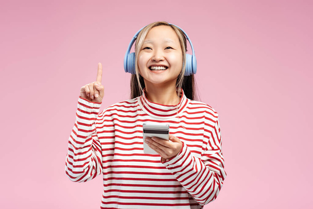 Glückliches asiatisches Mädchen posiert im Studio mit einem Handy, das Kopfhörer trägt und mit den Fingern nach oben zeigt, isoliert auf rosa. Kopierraum. Werbekonzept - Foto, Bild
