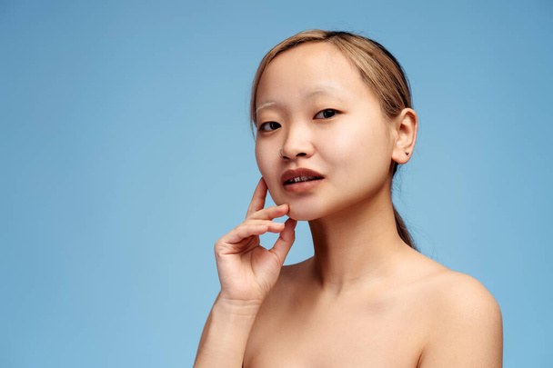Portrait of cute Asian girl posing with bare shoulders looking at camera, holding hand on cheek, isolated on blue. Beauty, body, skin care concept - Photo, Image