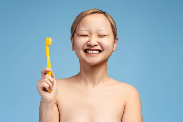 Mosolygó ázsiai lány portréja csukott szemmel, fogkefével pózolva, kék háttérrel. Fogászati ellátás fogalma - Fotó, kép