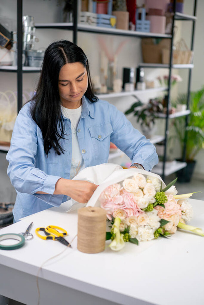 in een bloemenwinkel wikkelt een bloemist de afgewerkte tender in een witte papieren wikkel en bindt deze met een touw - Foto, afbeelding