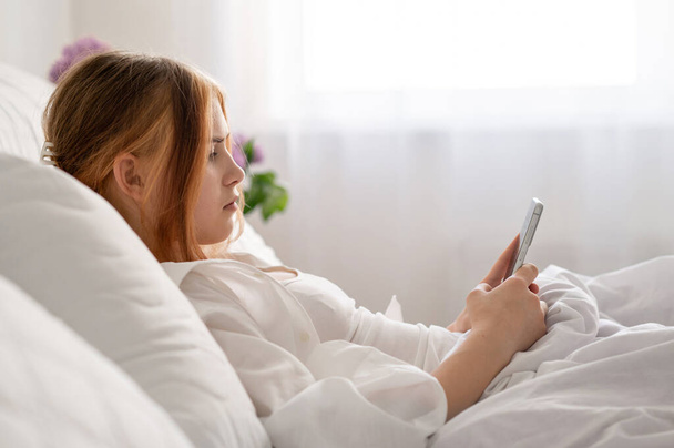 Teenage girl with mobile phone laying in bed, rest and leisure pastime, bad habits, youth gadget addiction. Neutral white airy background with soft natural light. - Photo, Image
