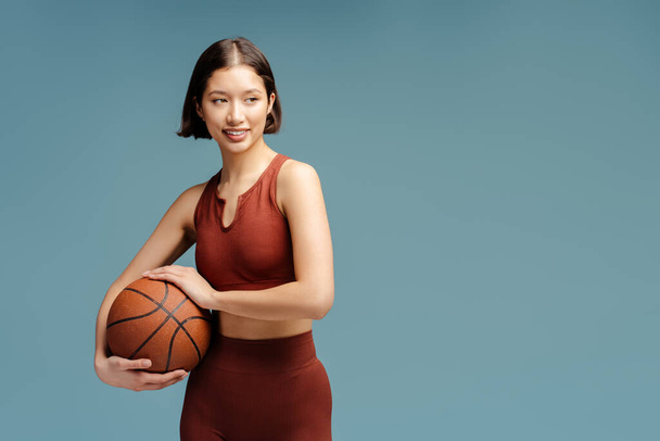 Retrato de una atractiva mujer sonriente jugando al baloncesto, sosteniendo la pelota mirando hacia otro lado, aislada sobre fondo azul. Deporte, afición, concepto de estilo de vida saludable - Foto, imagen