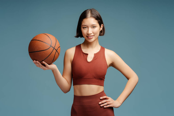 Portret van een mooie Aziatische vrouw die basketbal speelt, bal vasthoudend kijkend naar camera geïsoleerd op blauwe achtergrond, kopieer ruimte. Sport, hobby, gezond levensstijl concept - Foto, afbeelding