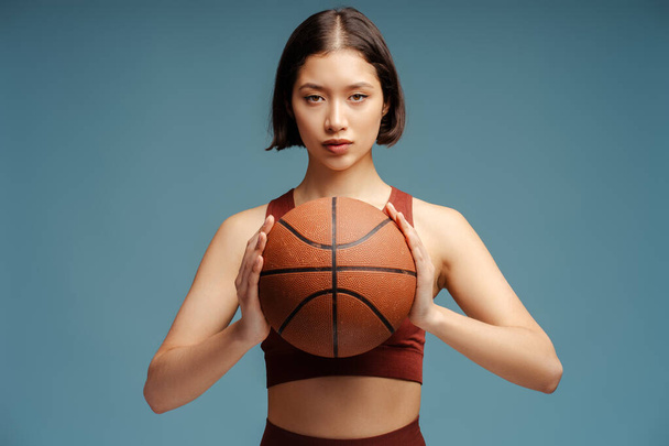 Σοβαρή γυναίκα που παίζει μπάσκετ, κρατάει μπάλα κοιτάζοντας την κάμερα, απομονωμένη σε μπλε φόντο. Έννοια αθλητικού ανταγωνισμού - Φωτογραφία, εικόνα