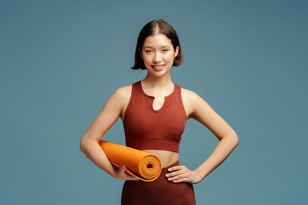 Confident young fit woman, with muscular strong aesthetic body, smiling, looking confidently at camera, carrying fitness yoga mat, isolated on blue background - Photo, Image
