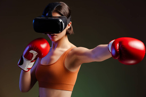 Портрет красивой серьезной женщины в очках виртуальной реальности, в боксёрских перчатках, делающей пунш на черном фоне  - Фото, изображение