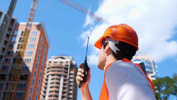 Constructor masculino usando walkie-talkie mientras habla en el sitio de construcción de pie cerca del edificio. Negocios, construcción, concepto de industria. Movimiento lento - Foto, imagen