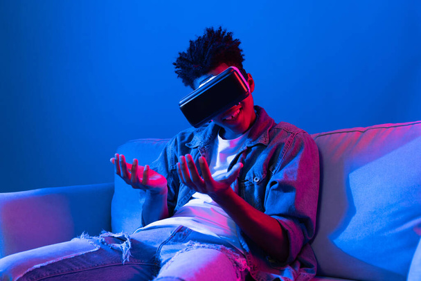 Junge afroamerikanisch aussehende VR genießen es, fantastische 3D-Zeigen interessante Objekttechnologie blaues Neonlicht Virtual Reality metaverse neue Welt im Wohnzimmer auf dem Sofa sitzen zu sehen. Beitrag. - Foto, Bild
