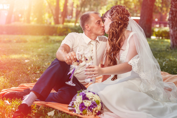 couple just married sitting in park green grass with bouquet of flowers and wine glasses - Photo, Image