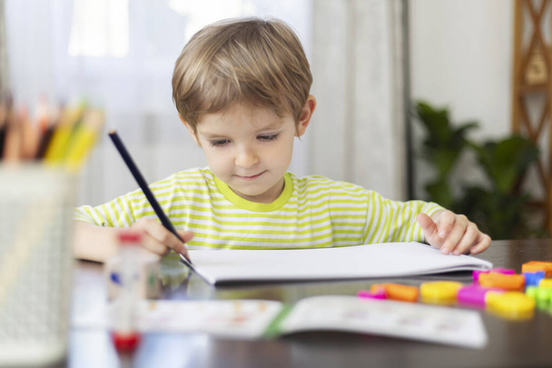 Egy fiatal fiú csíkos zöld ingben szorgalmasan ráfest egy fehér papírlapra, színes zsírkrétákkal körülvéve és fényes környezetben, ami fokozza a kreatív, játékos gyerekkor érzését. - Fotó, kép