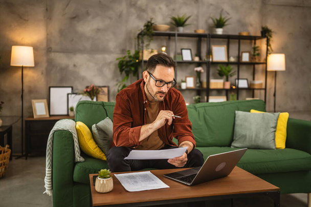 Homem caucasiano adulto com óculos trabalham em casa no laptop e mantêm documentos - Foto, Imagem