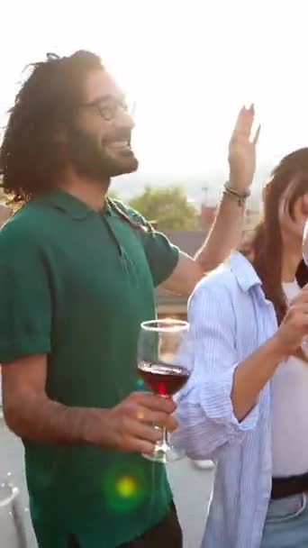 Verticale HD-video van jonge groep vrienden die plezier hebben met het roosteren van rode wijn terwijl ze samen dansen op het dak zomer zonsondergang feest. - Video