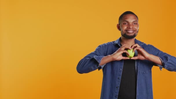 Afro-amerikan gevşemiş adam bir elmayla kalp şeklinde bir işaret veriyor ve stüdyoda yerel ürünleri tavsiye ediyor. Mutlu insan sağlıklı beslenmeyi ve kimyasalları besinsiz yemeyi sever. Kamera B. - Video, Çekim