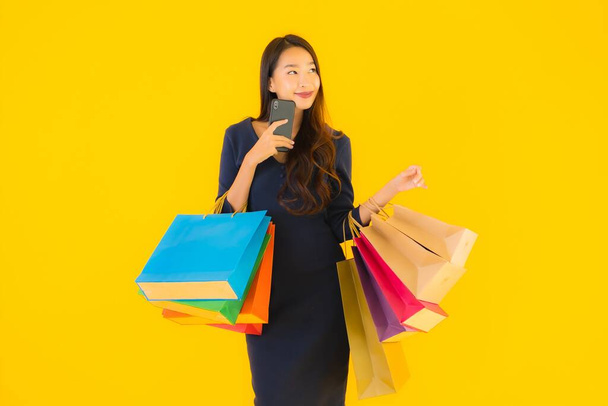 Portré gyönyörű fiatal ázsiai nő bevásárló táska hitelkártya és okos mobiltelefon sárga elszigetelt háttér - Fotó, kép