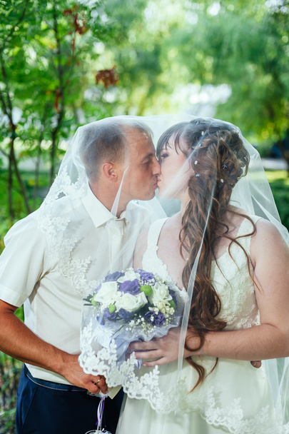 wedding couple kissing in green summer park. bride and groom kissing, standing together outdoors, hugging among green trees. Bride holding wedding bouquet of flowers - Valokuva, kuva