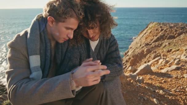 Békés pár ül a sziklán, kézen fogva az esti közelből. Vonzó férfi kötődik egy gyönyörű nőhöz a tengerparton. Gyengéd családi pár - Felvétel, videó