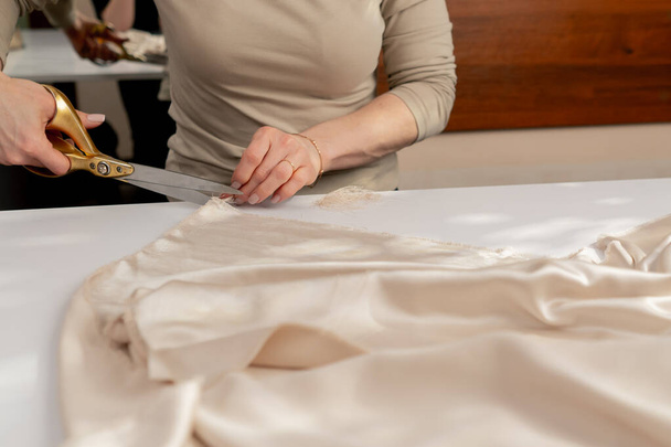 close up Una costurera profesional corta tela de seda crema con tijeras para un producto personalizado vestido - Foto, Imagen