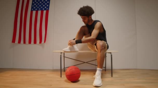 Dolly-in slow mo shot di pallacanestro maschile Biracial che lega i lacci delle scarpe su una panchina di legno nello spogliatoio con bandiera statunitense appesa al muro - Filmati, video