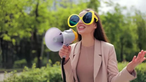 Emotional caucasian woman walking through roadway with white loudspeaker in hands. Confident female wearing big glasses with blue lenses and elegant jacket attracting attention of people on street. - Footage, Video