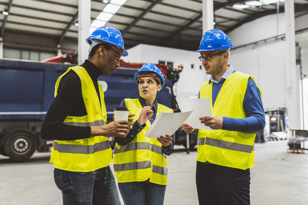 A biztonsági felszerelésben dolgozó mérnökök sokszínű csoportja tervekről és dokumentumokról tárgyal egy ipari létesítményben. - Fotó, kép