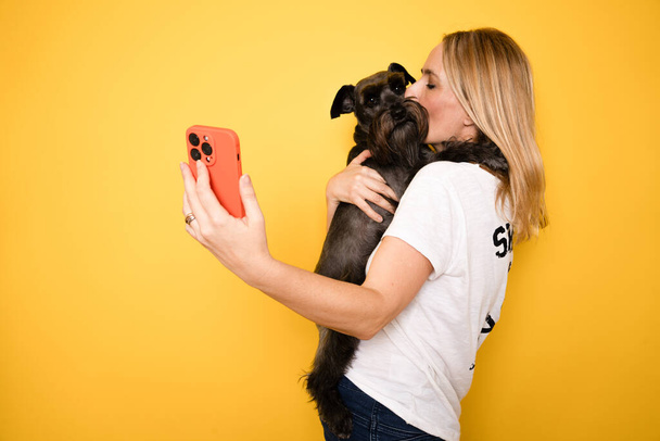 Крупним планом молода жінка-власник зі своєю найкращою подругою собакою-шнаузером носить повсякденний одяг робити селфі, знятий на мобільний телефон на жовтому тлі - Фото, зображення