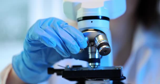 Científico utiliza microscopio para examinar la muestra. Asistente de laboratorio ajusta lente del microscopio en laboratorio - Imágenes, Vídeo
