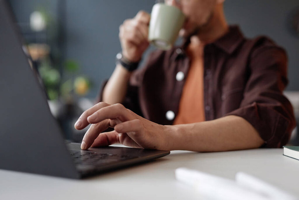 Közelkép felismerhetetlen férfi gépelés laptop billentyűzeten, miközben dolgozik az íróasztalnál, és iszik kávé másolási hely - Fotó, kép