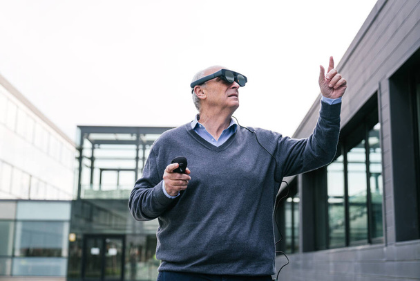 Vanhempi johtaja puserossa, joka on vuorovaikutuksessa virtuaalitodellisuuslasien kanssa seisoessaan modernin toimistorakennuksen ulkopuolella. - Valokuva, kuva