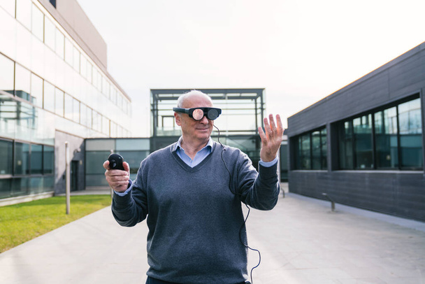 Un cadre supérieur portant un pull s'engage avec des lunettes de réalité virtuelle, interagissant avec des éléments virtuels à l'extérieur d'un immeuble de bureaux moderne - Photo, image