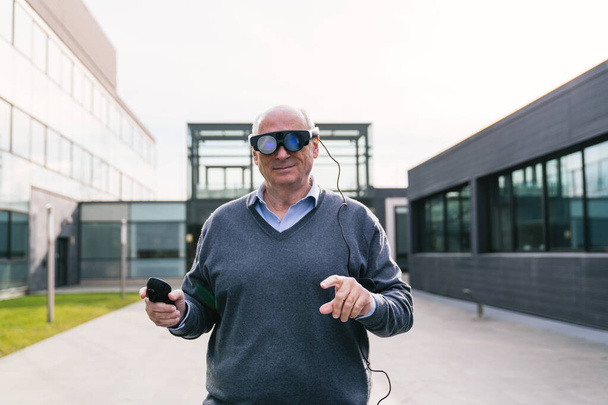 Un hombre de negocios senior que usa gafas de realidad virtual y usa un controlador, parado fuera de un moderno edificio de oficinas. - Foto, Imagen