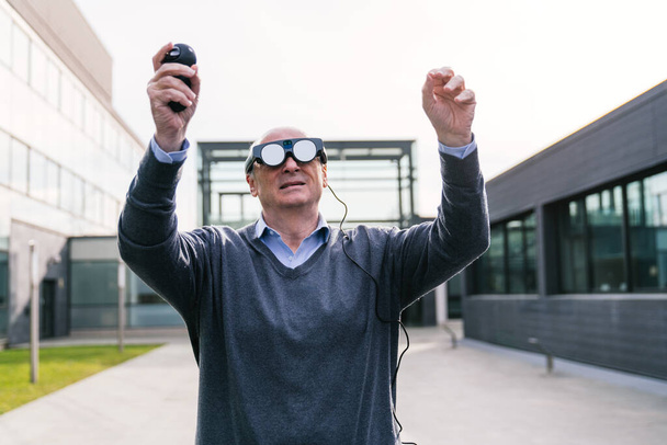 Um empresário sênior usando óculos de realidade virtual e um controlador, engajando-se em uma experiência interativa fora de um prédio de escritórios moderno. - Foto, Imagem
