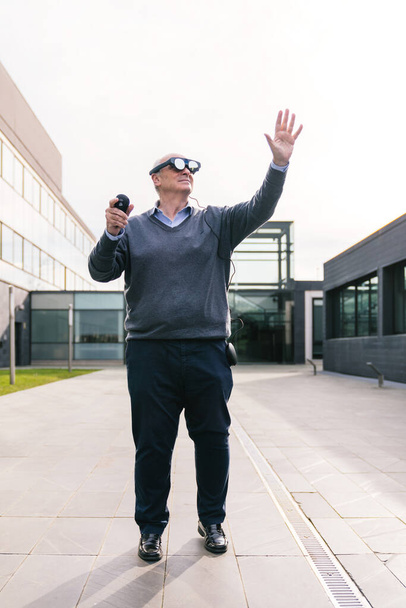 Cadres supérieurs utilisant des lunettes de réalité virtuelle et un contrôleur, s'engageant avec des éléments virtuels à l'extérieur d'un immeuble de bureaux moderne. - Photo, image