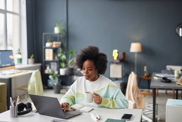 Широкий угол обзора афроамериканской женщины, работающей в одиночку за столом в офисе против серых стен копировального пространства - Фото, изображение